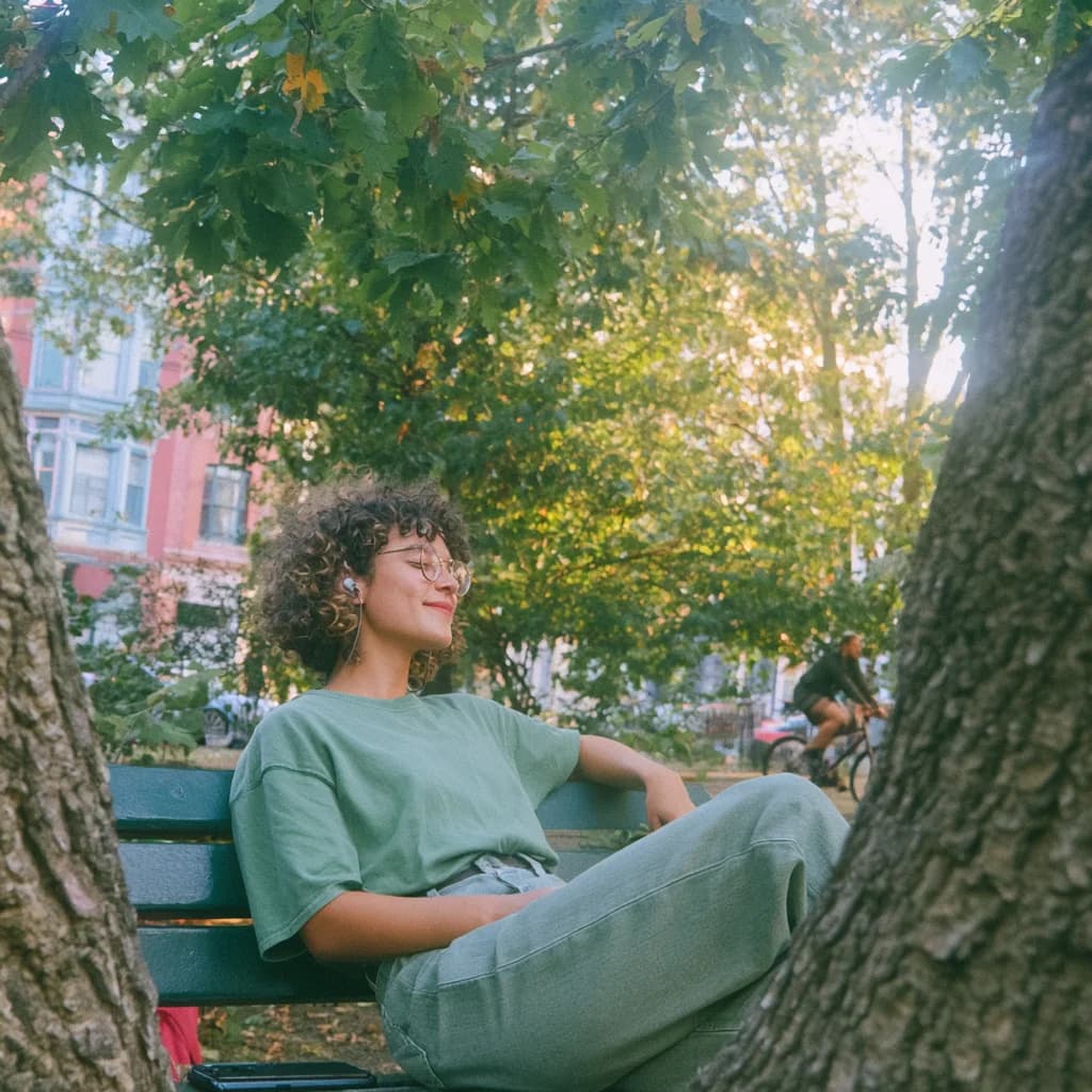 A person listening through earbuds on a bench beneath trees